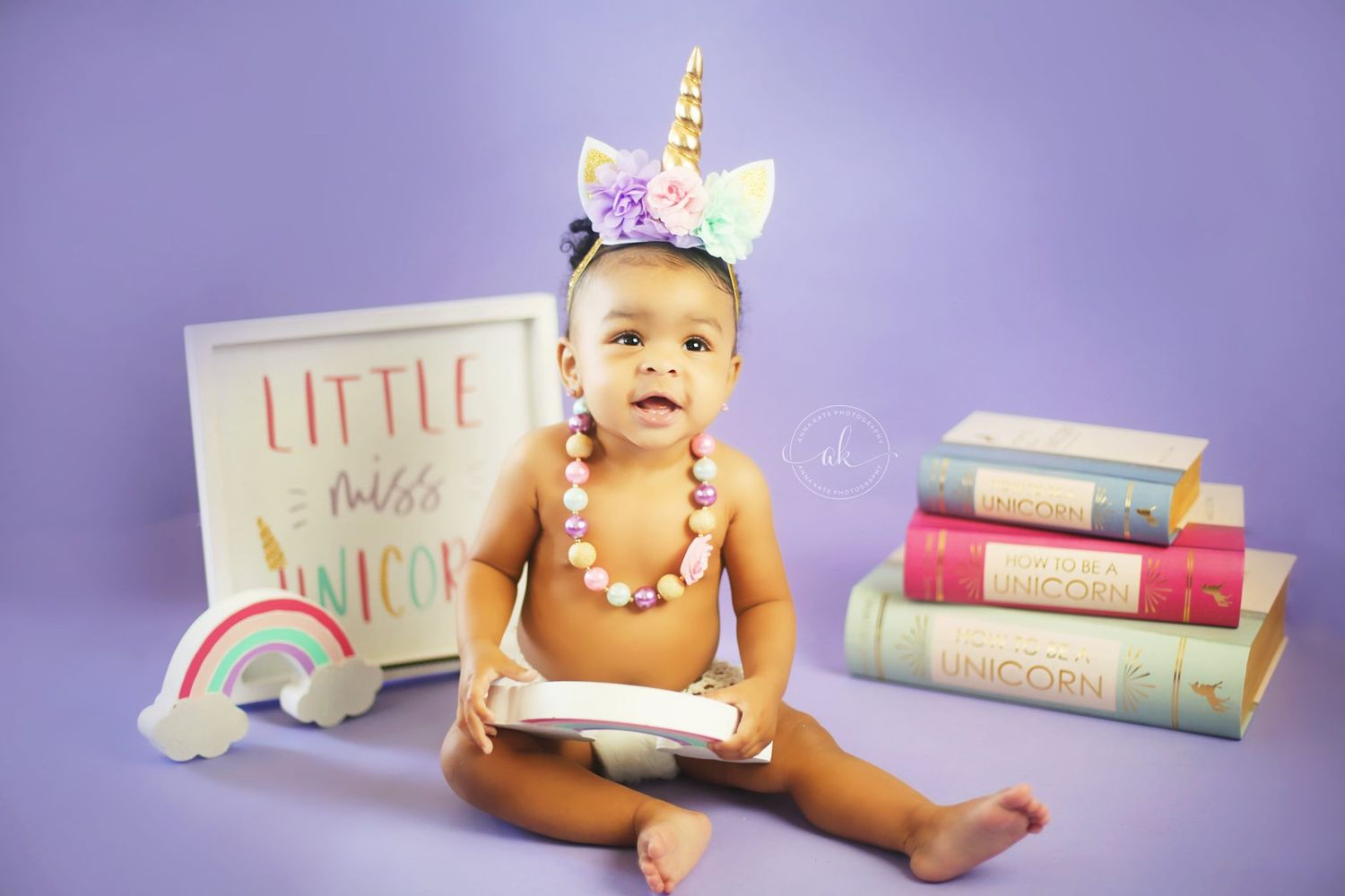 Baby photo with unicorn headband, books and rainbow decorations against purple background.
