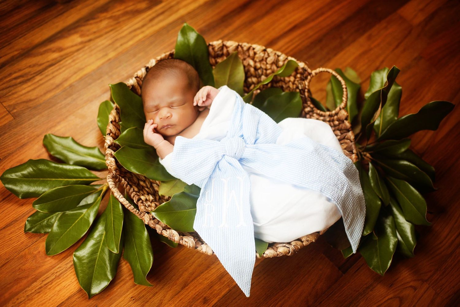 Sleeping newborn in white dress laying in woven basket decorated with green leaves on wooden floor.