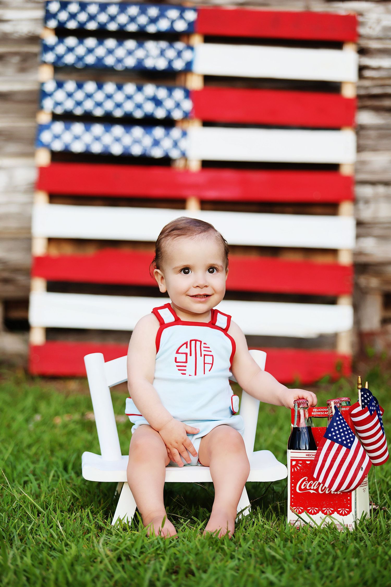 A patriotic holiday setup with wooden American flag decor and Coca-Cola bottles on green grass.