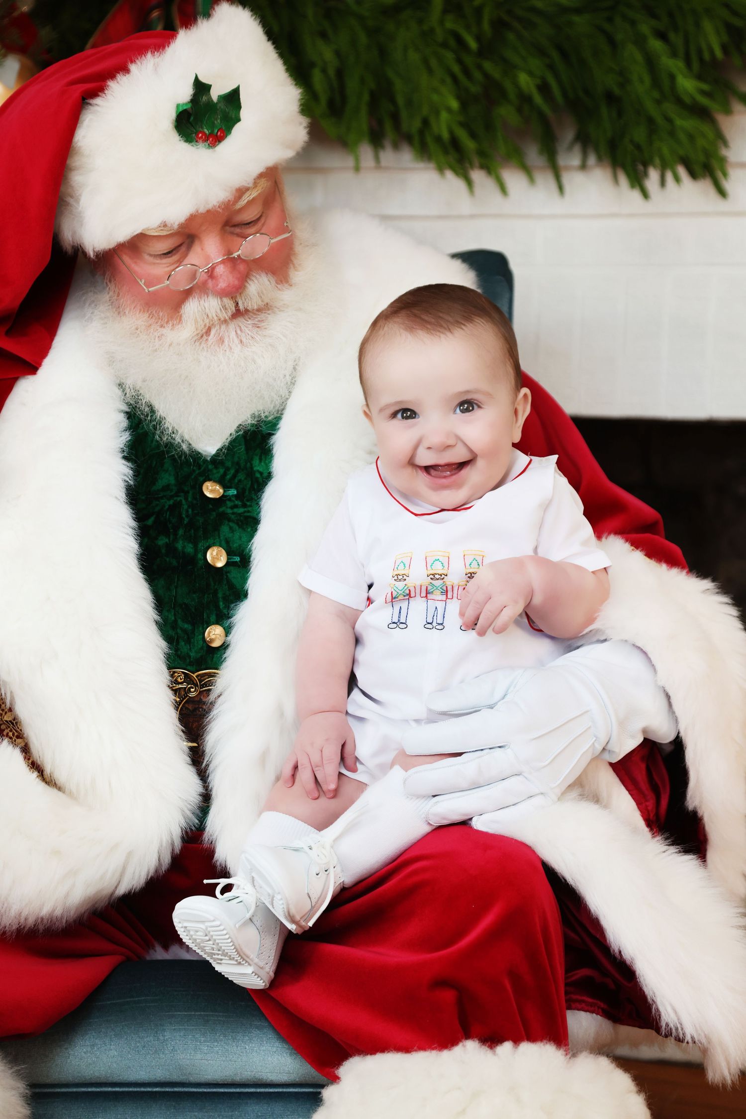 Holiday photo series shows baby in white outfit sitting with Santa Claus in traditional red suit and white beard.