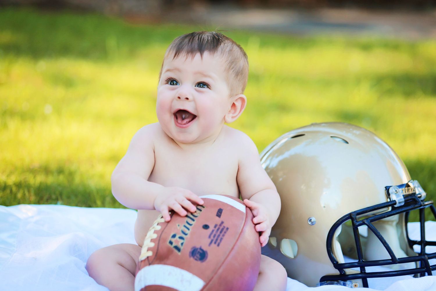 Baby on blanket outdoors with football and helmet props.