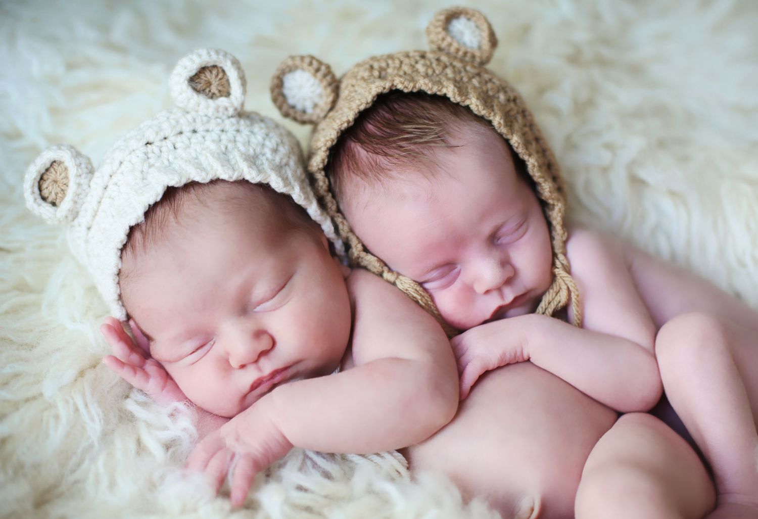 Sleeping newborns wearing adorable bear-ear bonnets on soft white fabric.