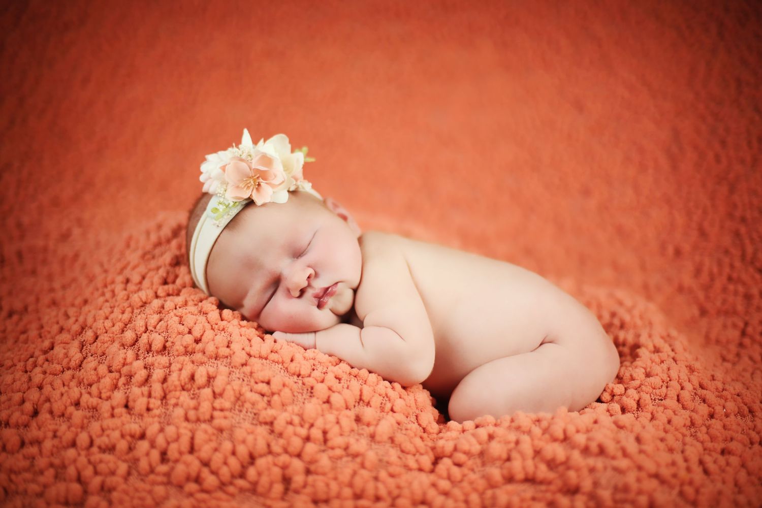 Sleeping newborn on orange textured blanket wearing delicate floral headband.