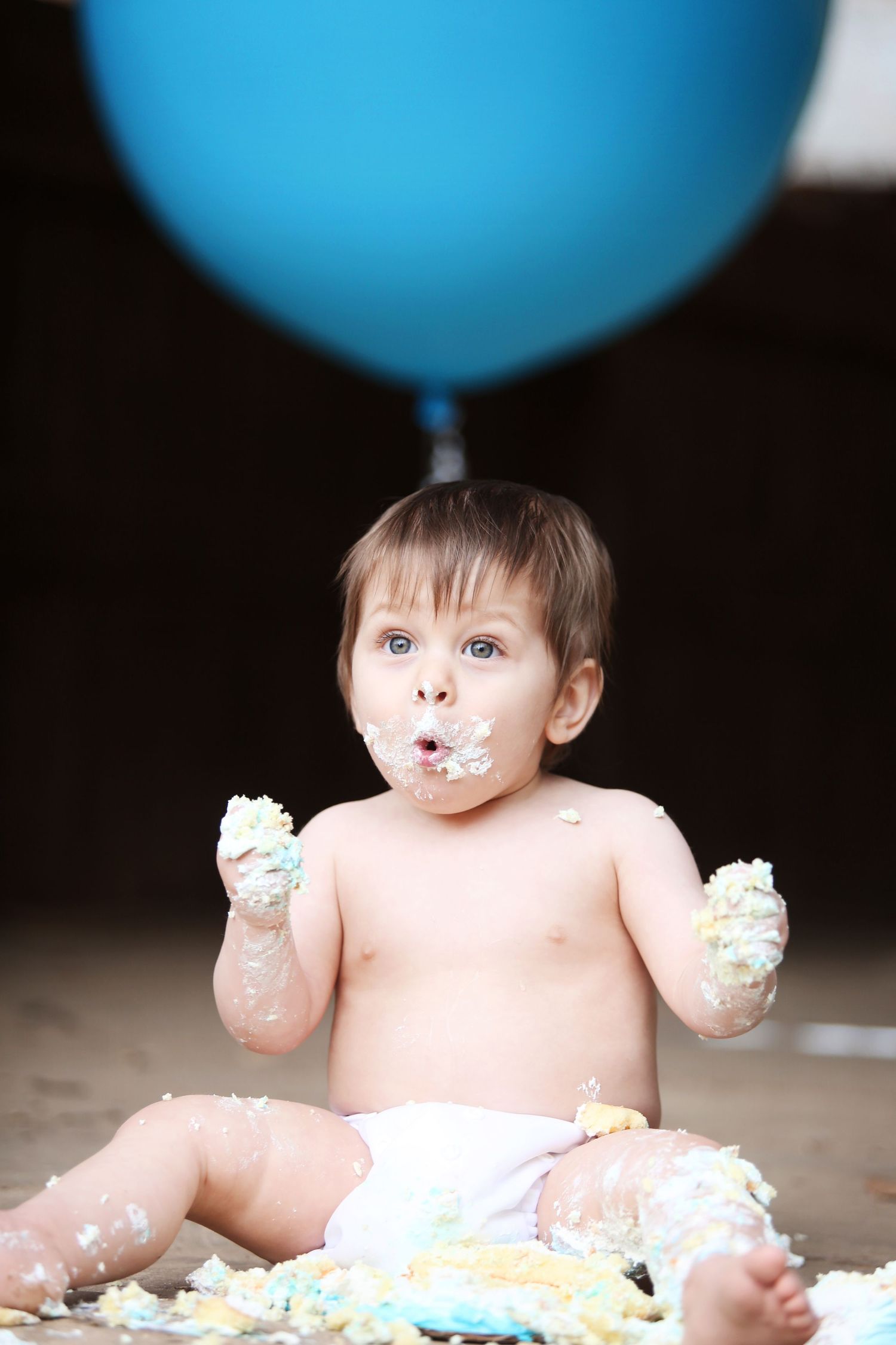 Baby covered in cake frosting with large blue balloon overhead.