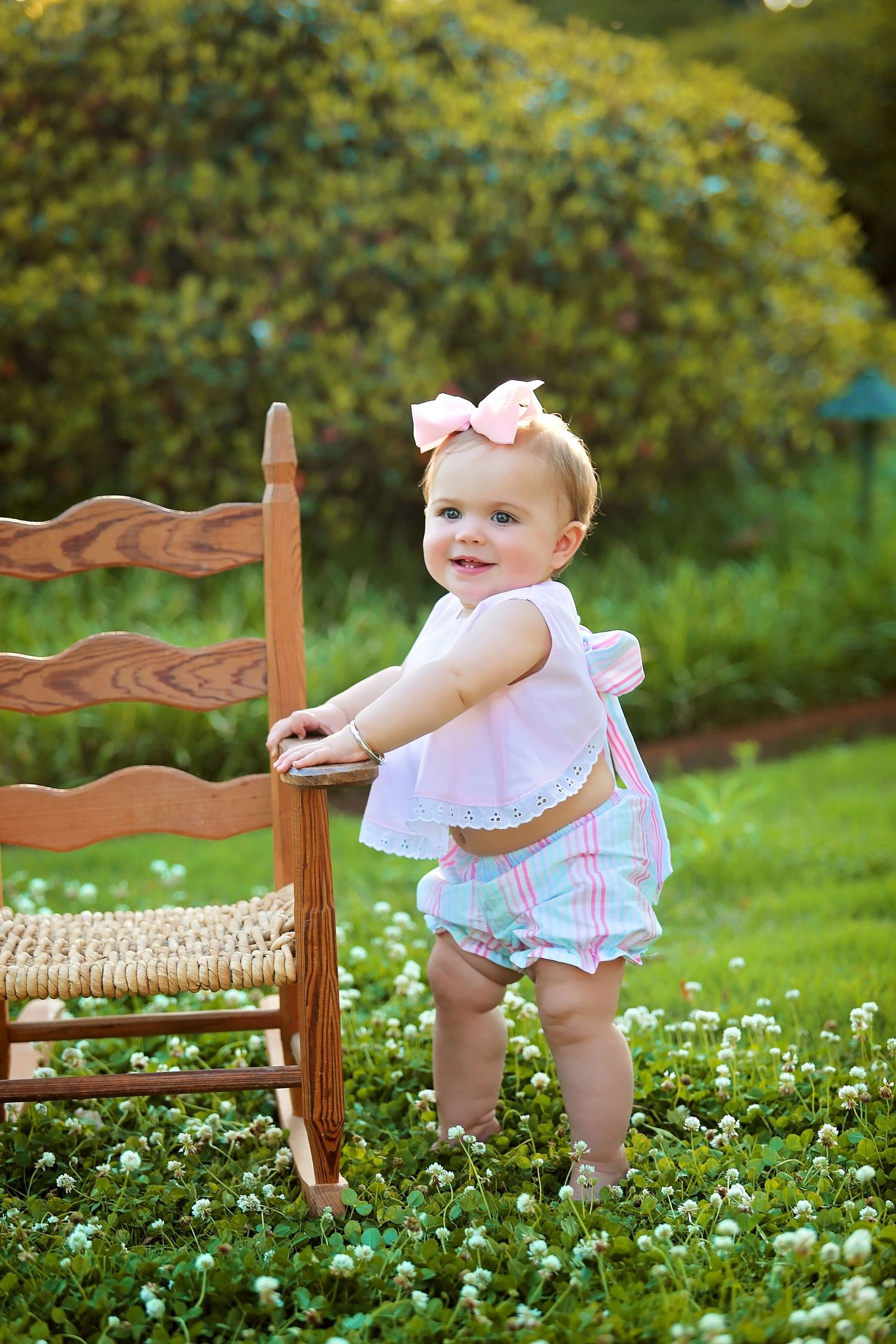 Small child in pink and white outfit standing next to wooden chair in garden with blooming flowers.