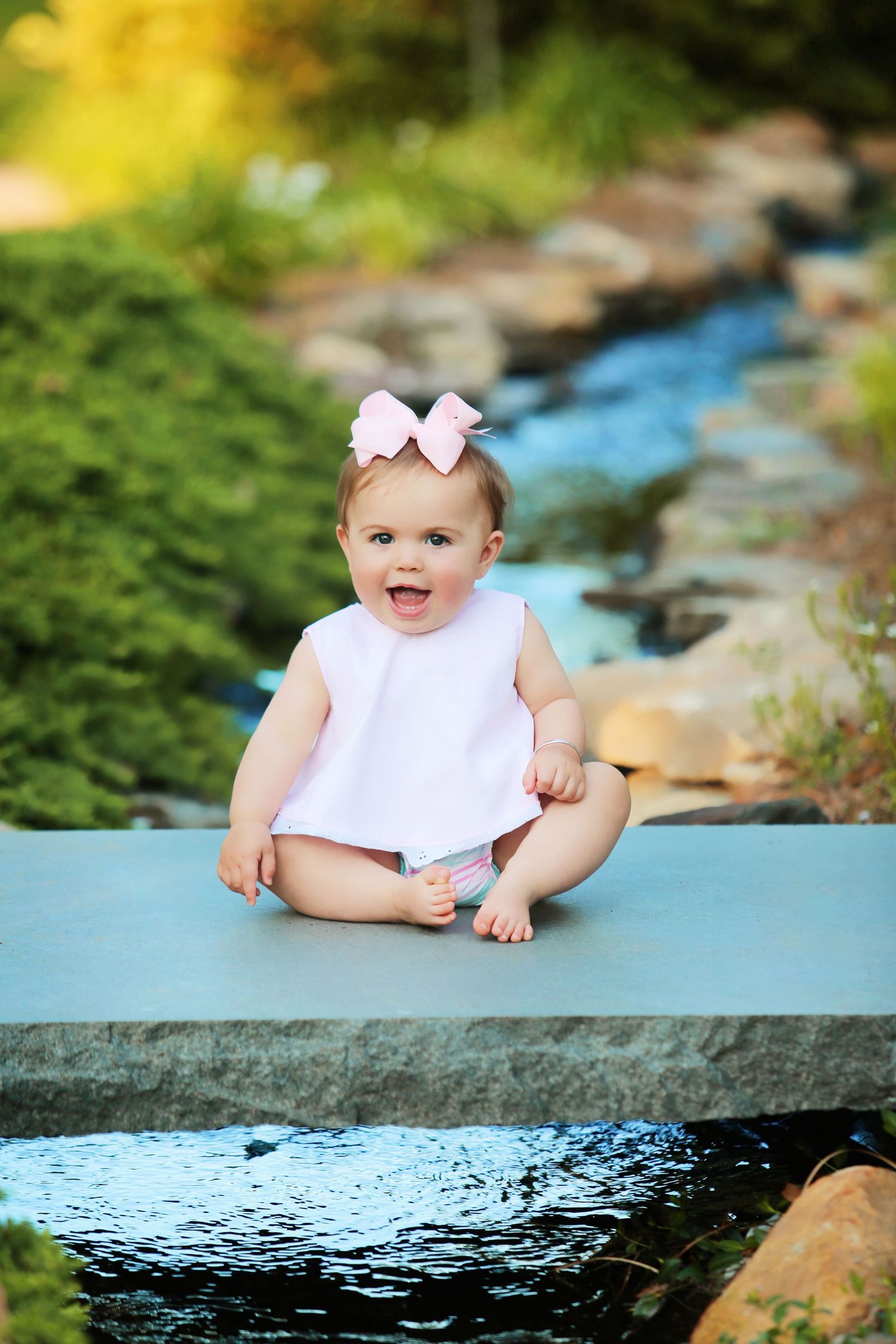 Baby in white dress with pink bow sits near flowing water feature.