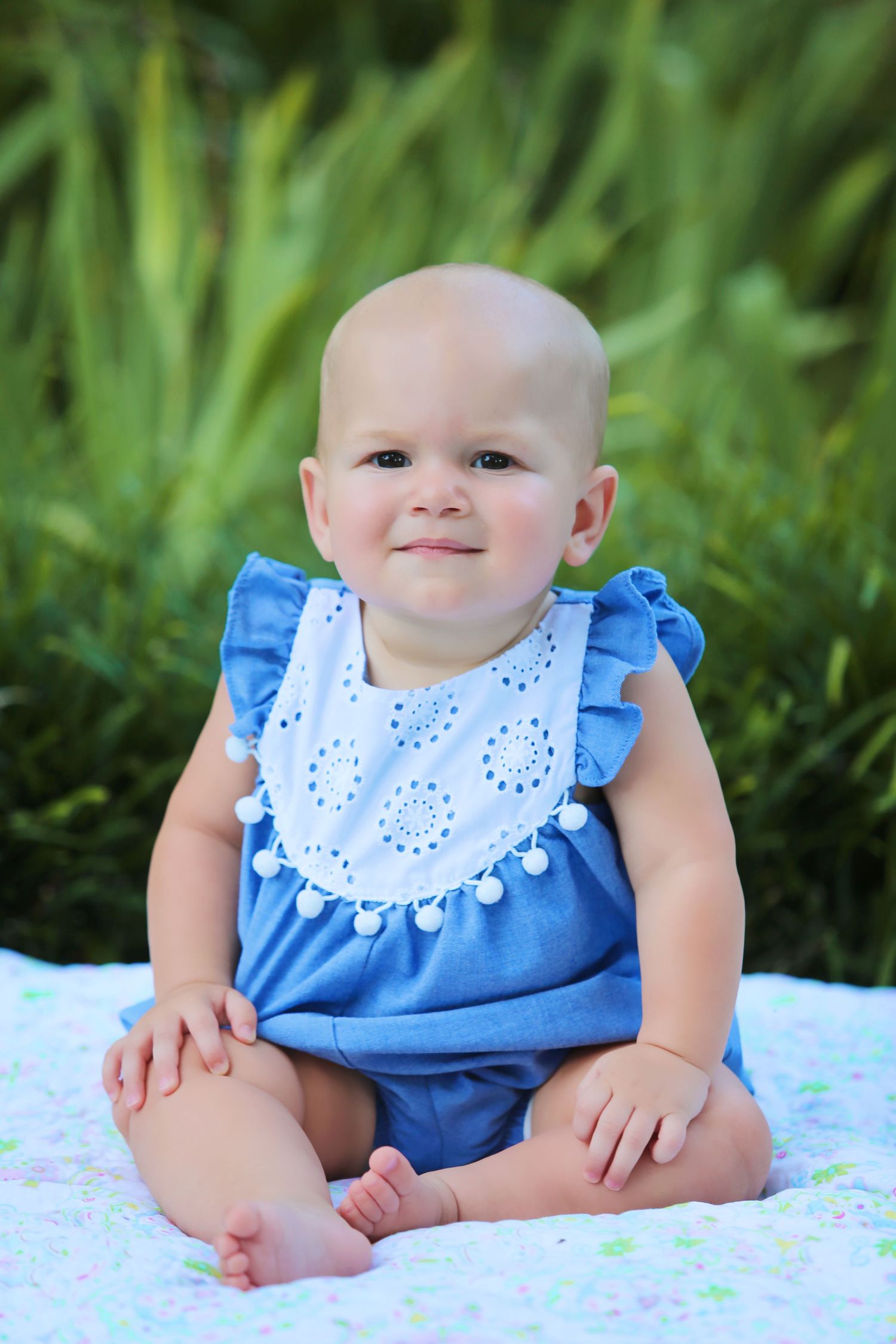 Baby wearing blue and white ruffle dress sits on blanket in grass.