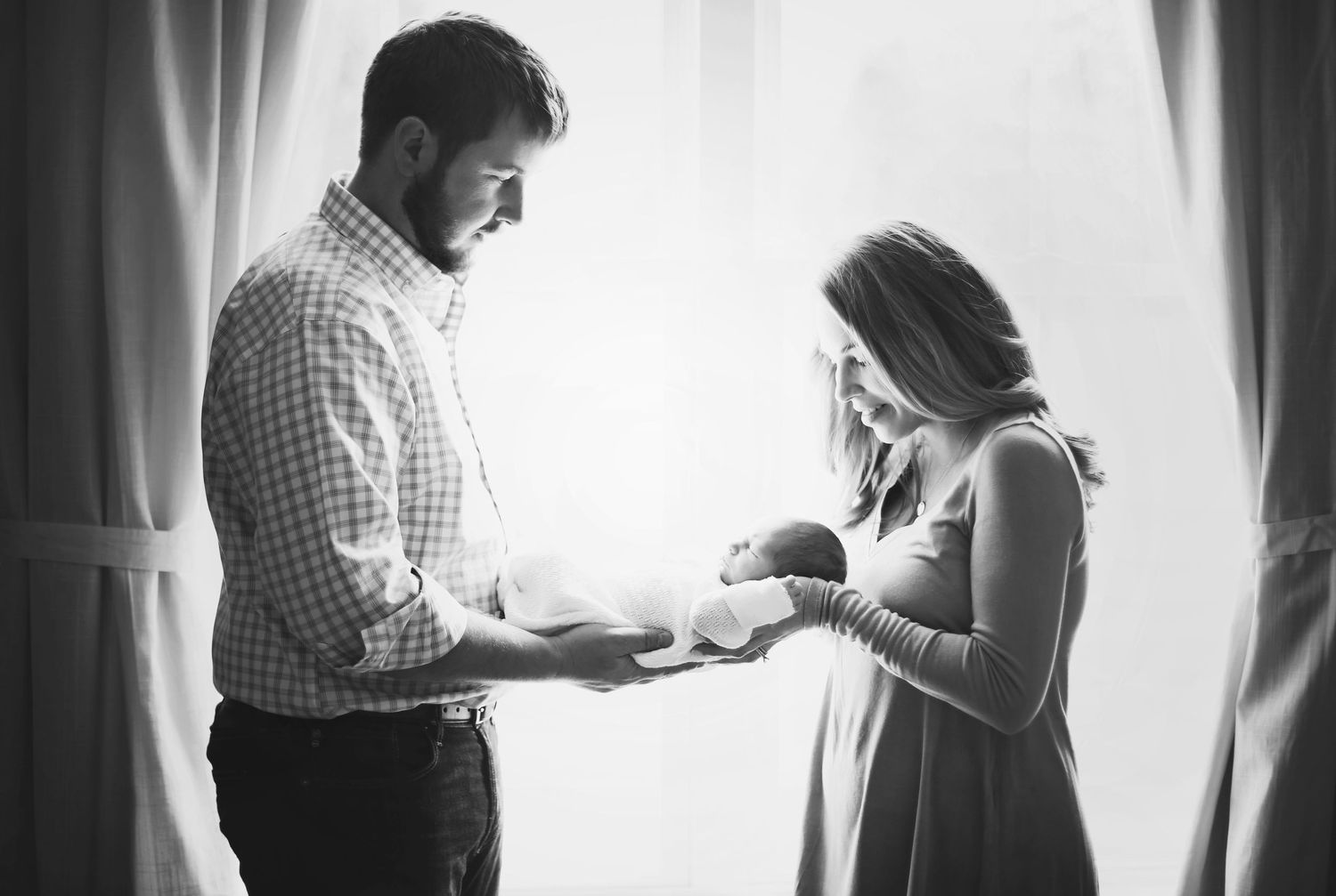 Black and white photo of couple holding newborn by window.