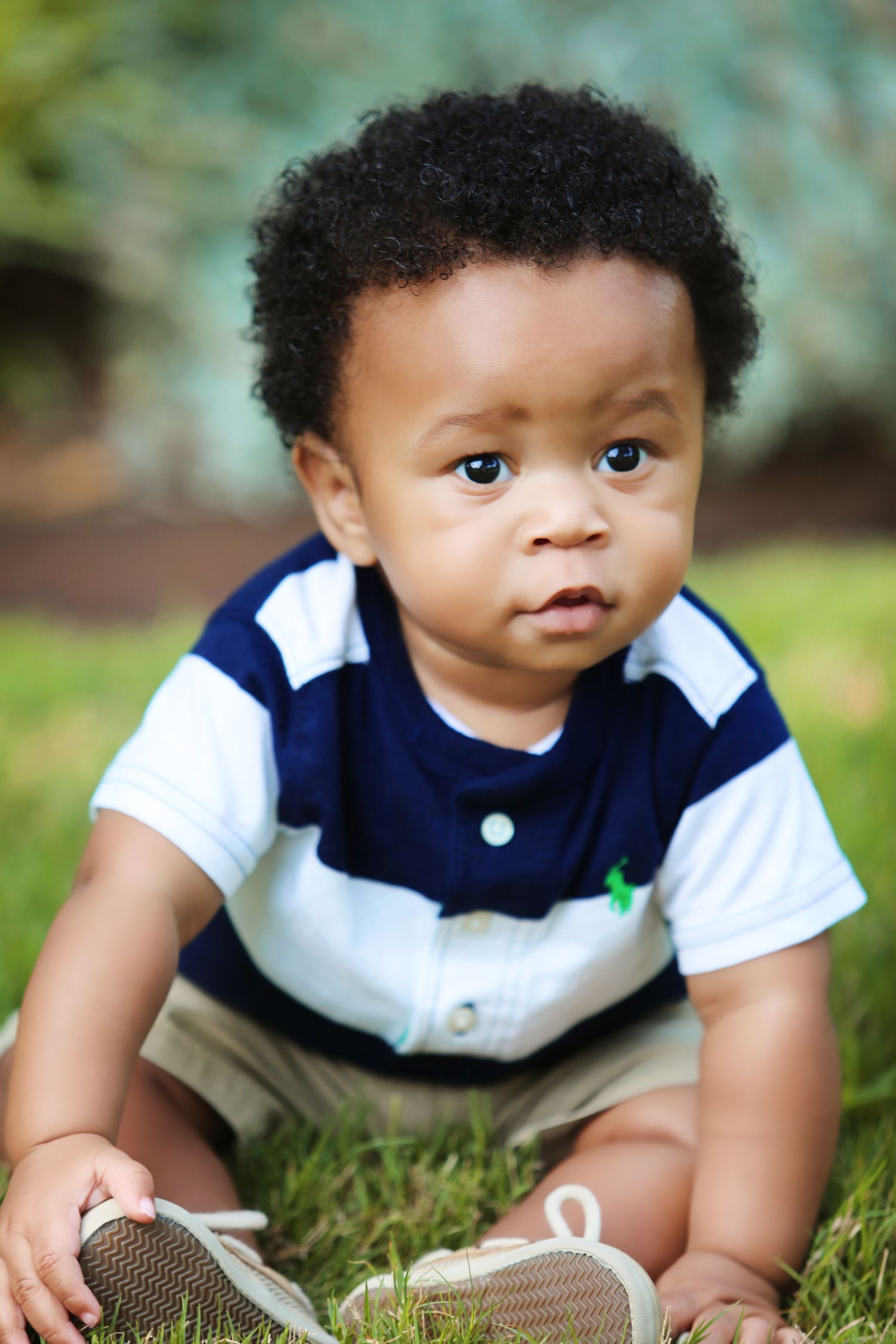 Baby in navy striped outfit sitting on grass wearing sneakers.