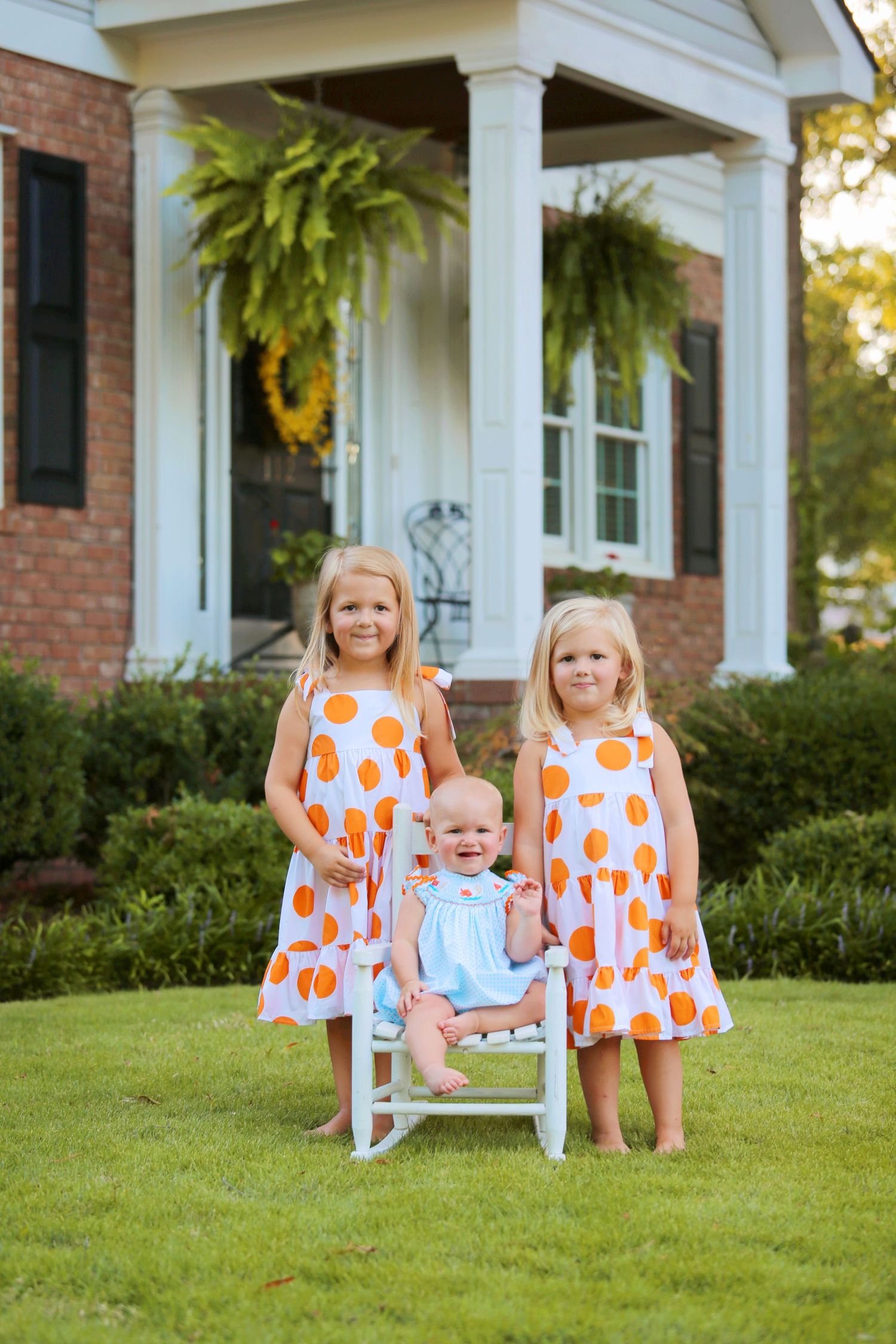 Three children in matching orange polka dot dresses stand in front of house.