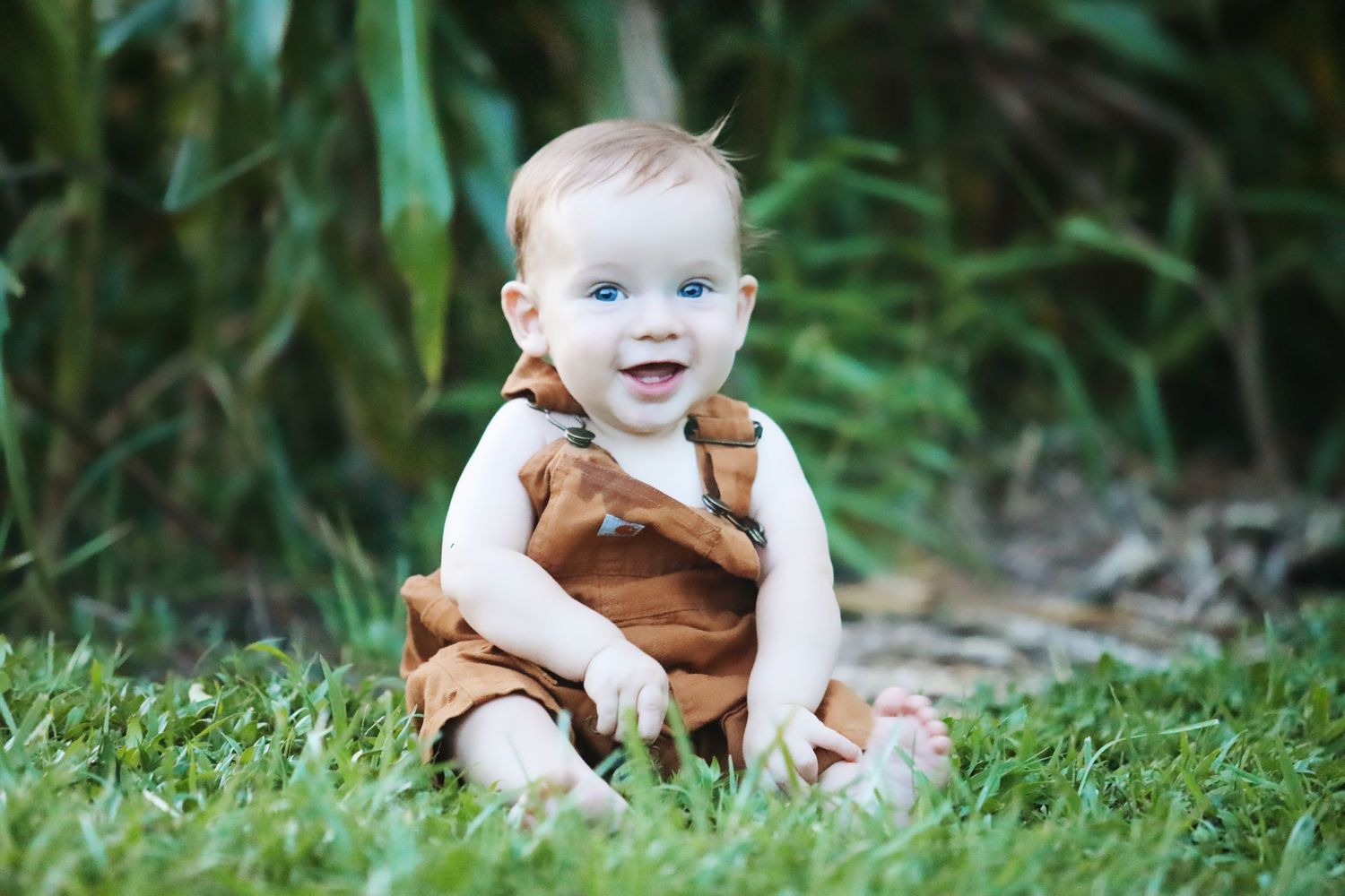 Happy baby in brown romper sitting on grass outdoors.