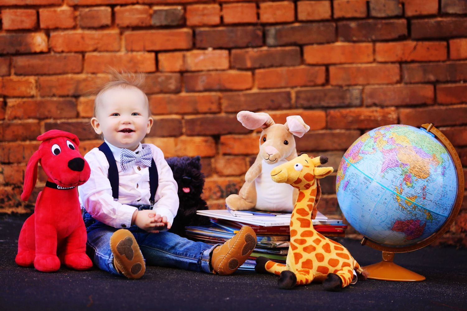Baby sitting with stuffed animals, globe and toys against brick wall background.