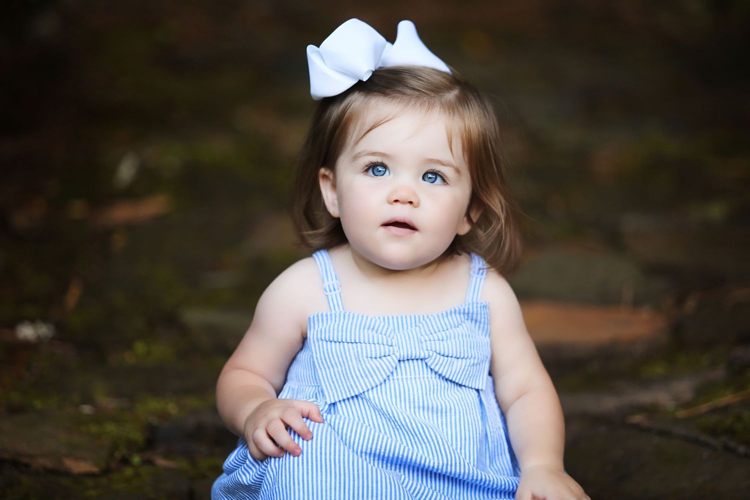 Toddler in blue striped dress with white bow sits outdoors.