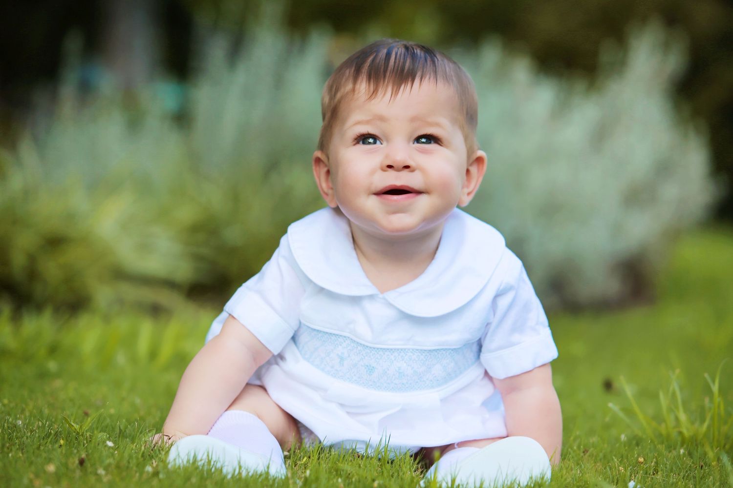 Baby wearing white outfit sitting in grass and smiling.