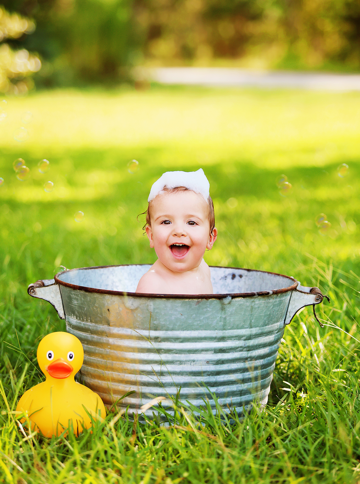 A smiley baby gets ready to splash in a vintage-style metal washtub outdoors with a yellow rubber duck toy beside it.