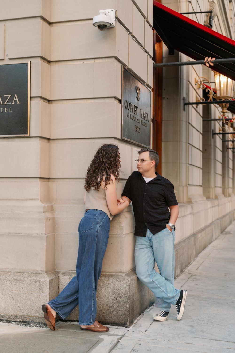 Couple poses casually along an urban building exterior with large windows.