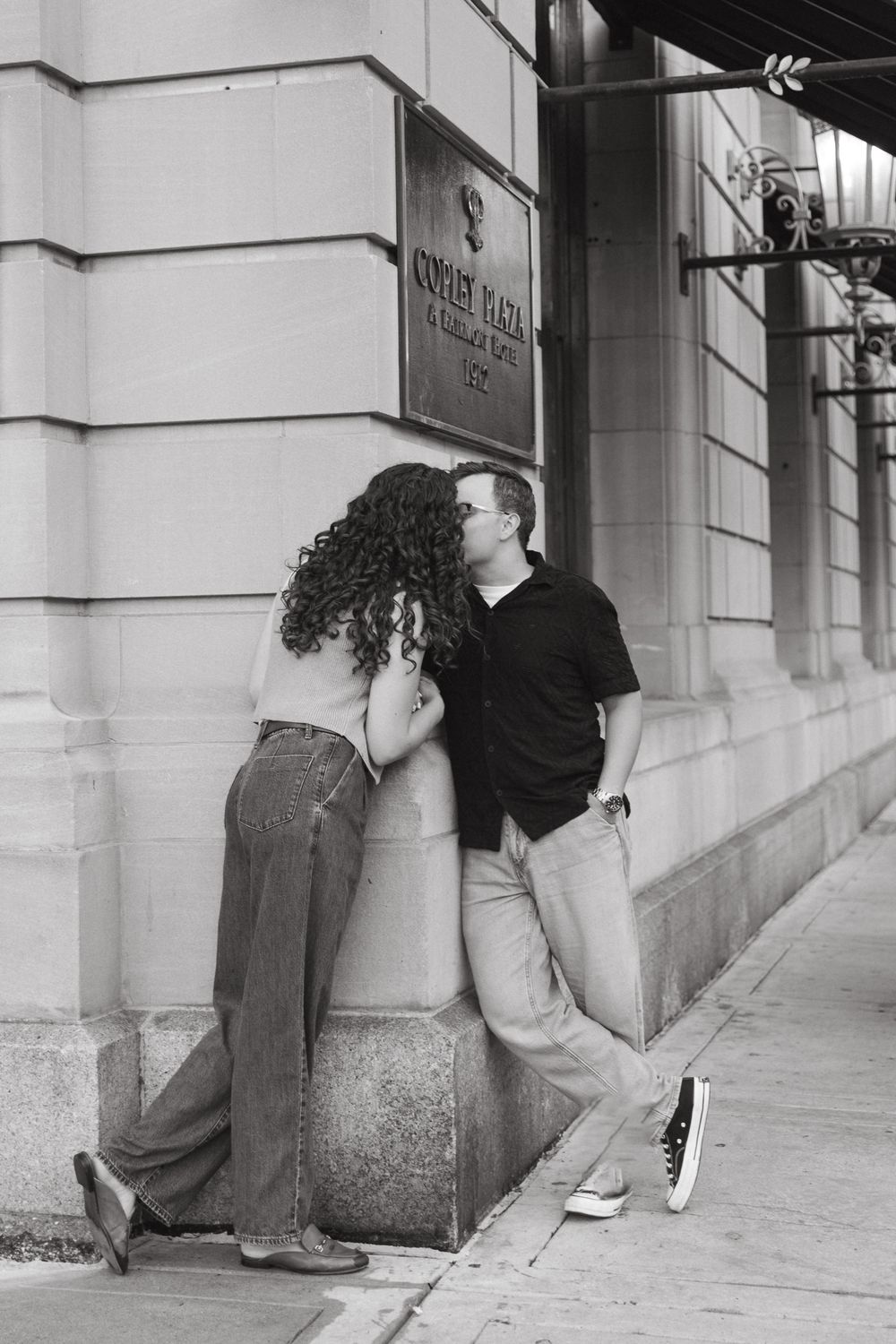 Black and white photo of a couple being affectionate by a building wall.