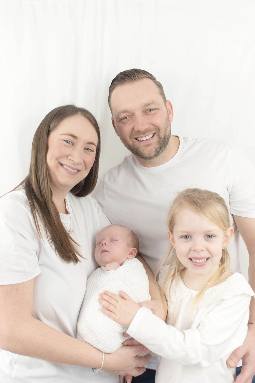 Family wearing white poses together for bright studio portraits with their newborn.
