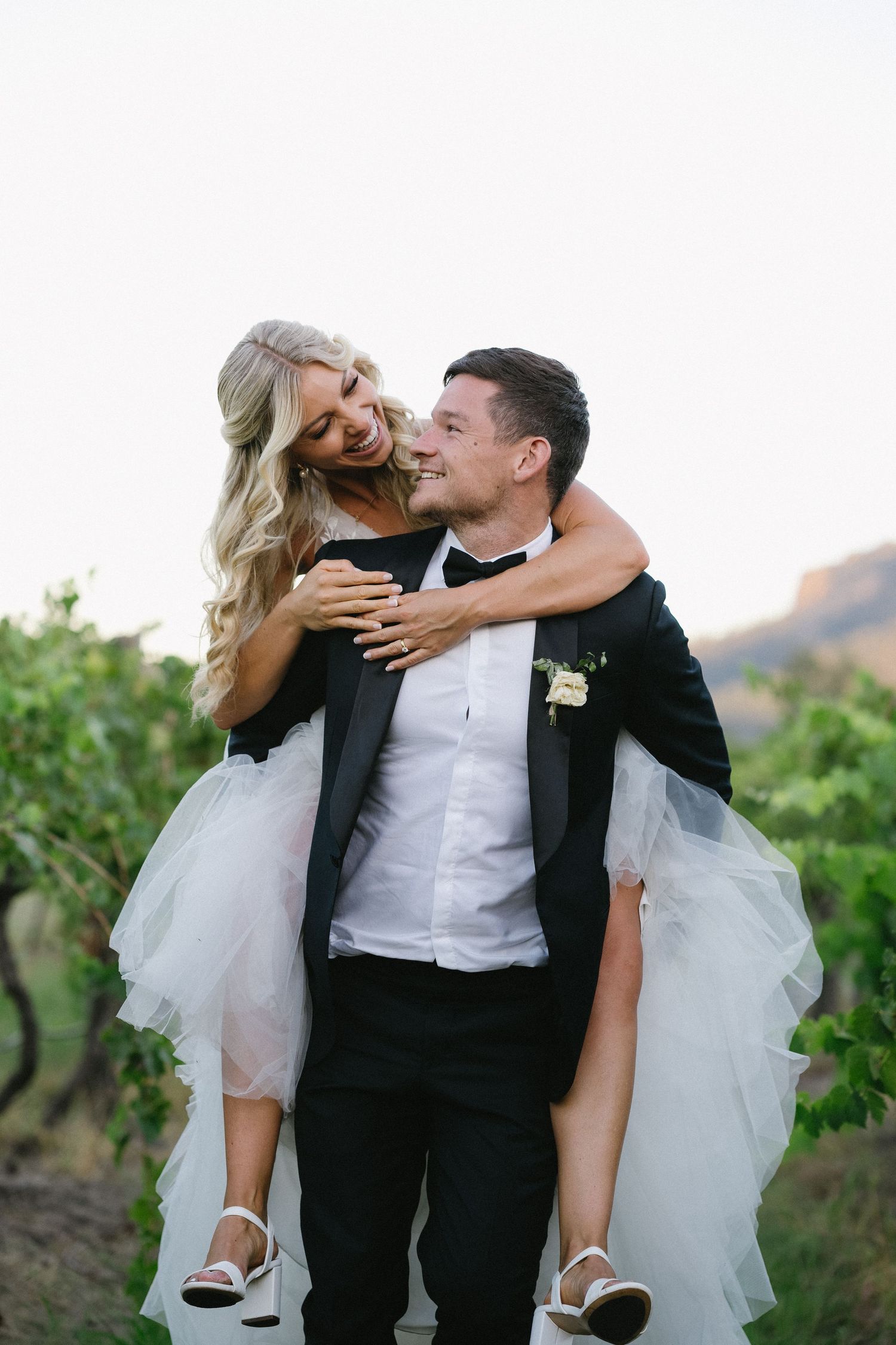 Groom gives bride playful piggyback ride through vineyard in wedding attire.