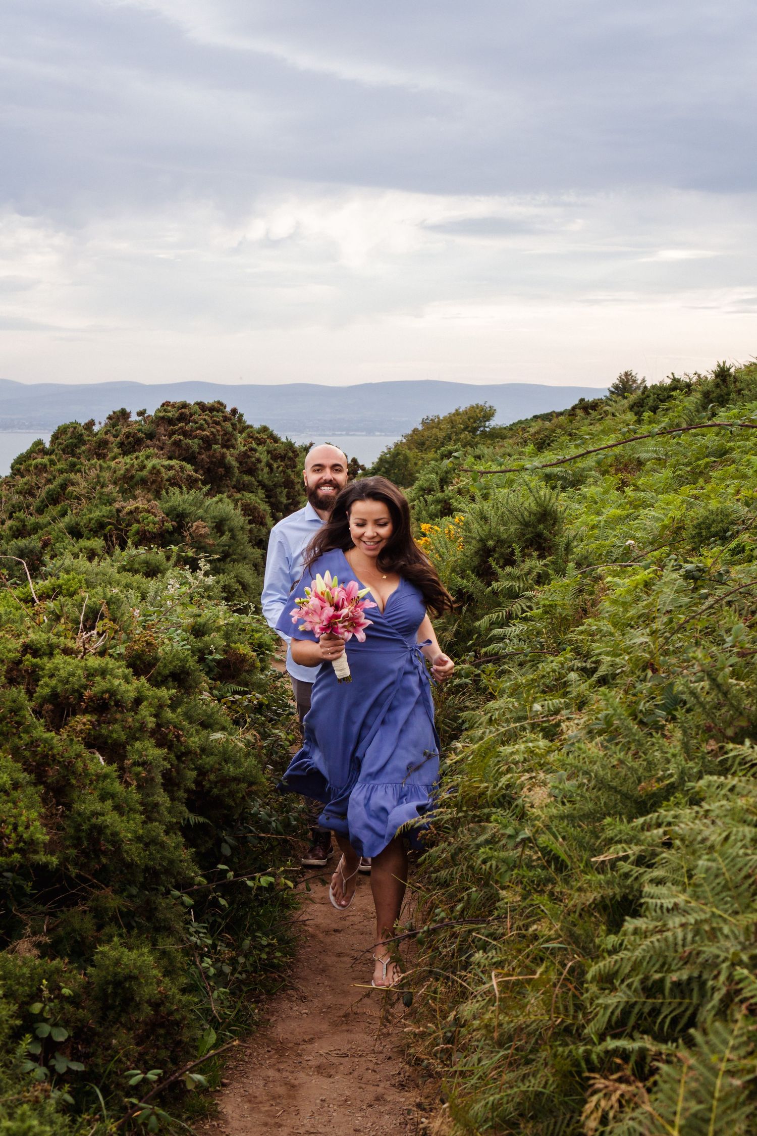 couple running towards the camera at howth cliff she's on a blue dress and he's on a blue shirt