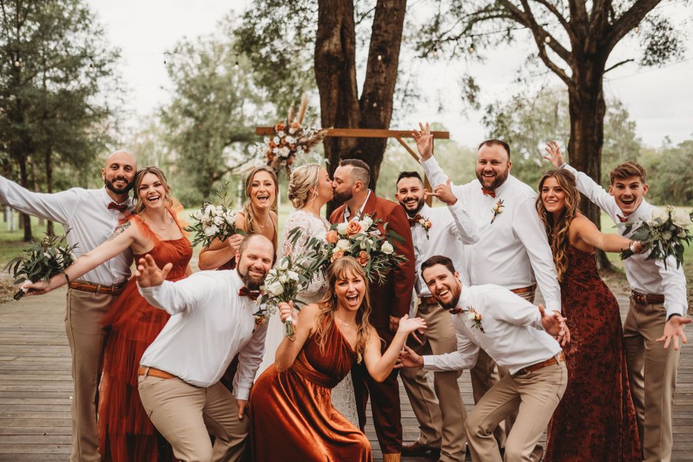 Wedding party celebrates with fun poses and expressions on wooden deck in rustic outdoor setting.