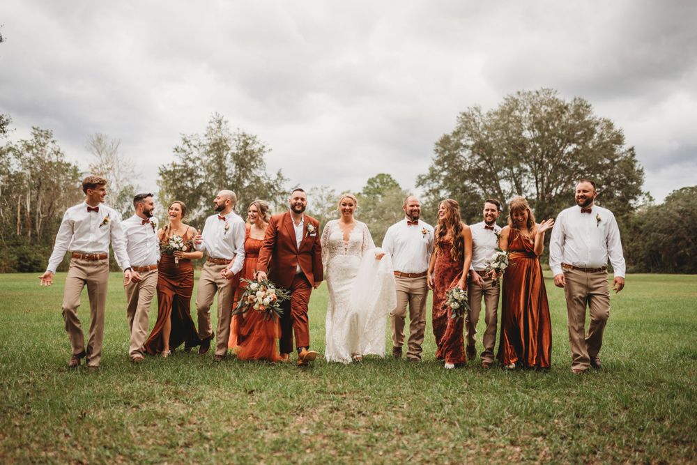 Wedding party poses on grass with burgundy and white color scheme under cloudy sky.
