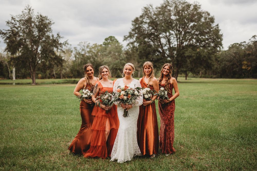Bride and bridesmaids in rust-colored dresses pose together holding bouquets in open field.