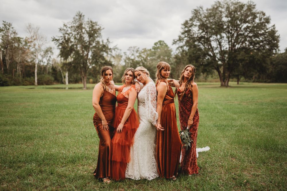 Bride in white gown poses with bridesmaids wearing rust-colored dresses in grassy field.