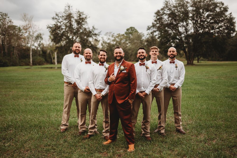 Wedding party in white shirts and khakis stands outdoors with one person wearing a rust-colored suit.