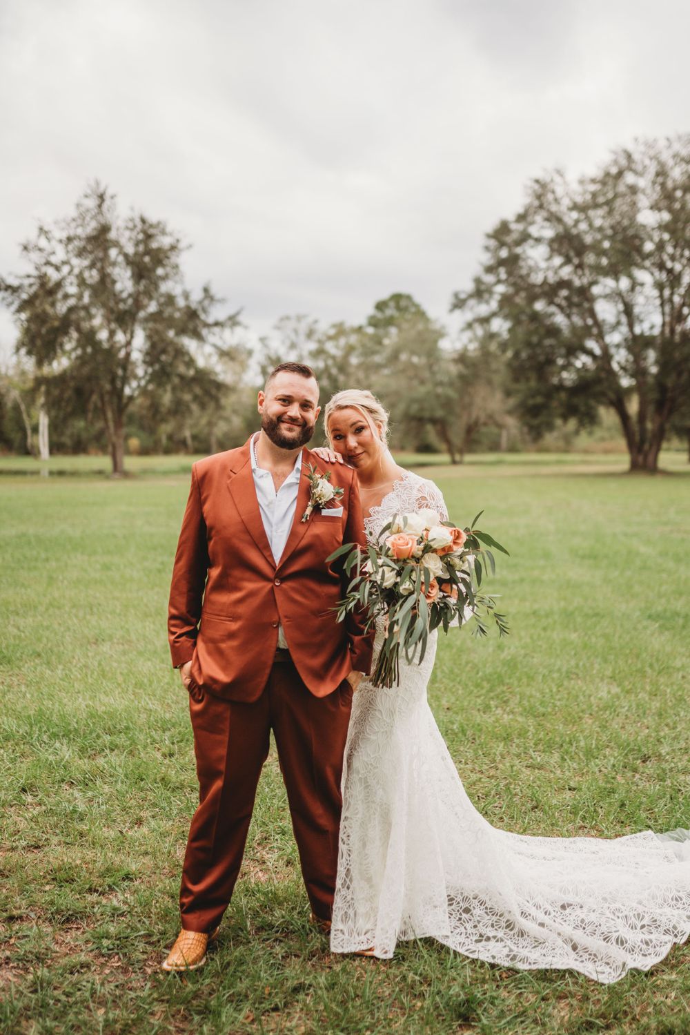 Newlyweds pose together outdoors, featuring a rust-colored suit and lace wedding gown with trailing veil.