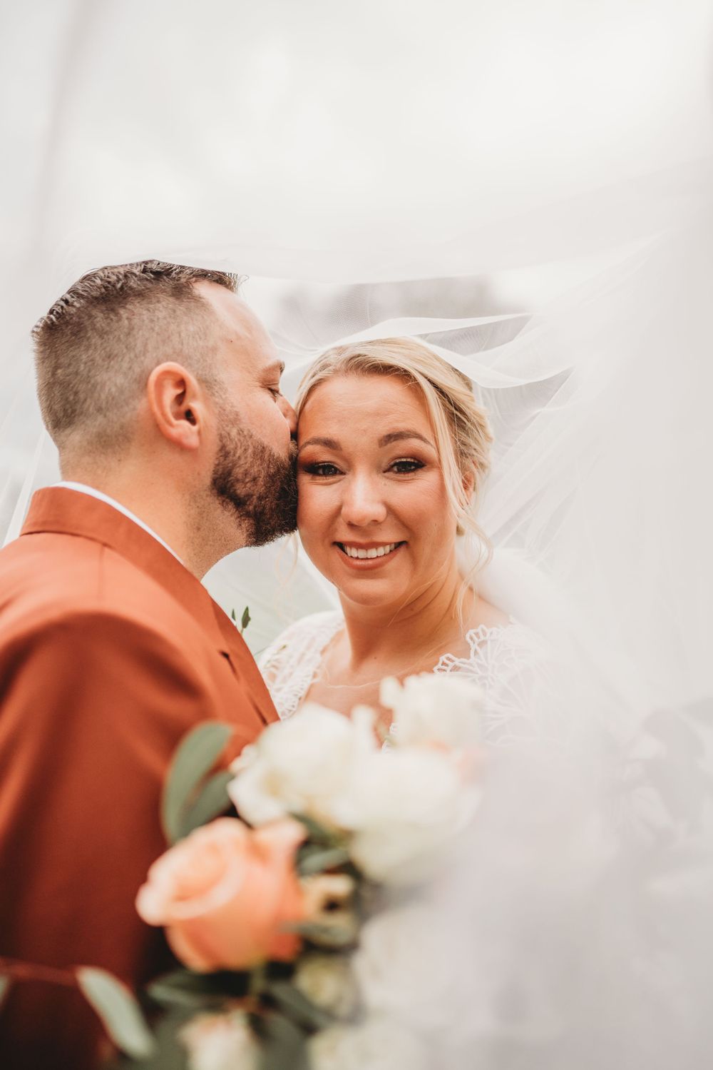 Intimate wedding moment captured with rust suit and flowing veil against bright background.