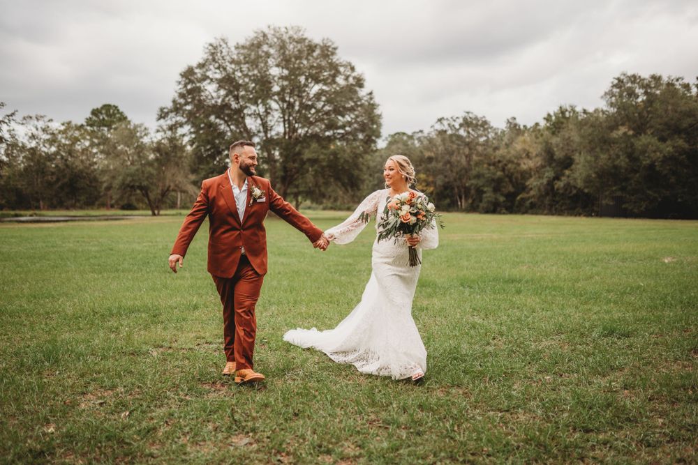 Couple walks hand in hand across grassy field in rust suit and lace wedding dress.