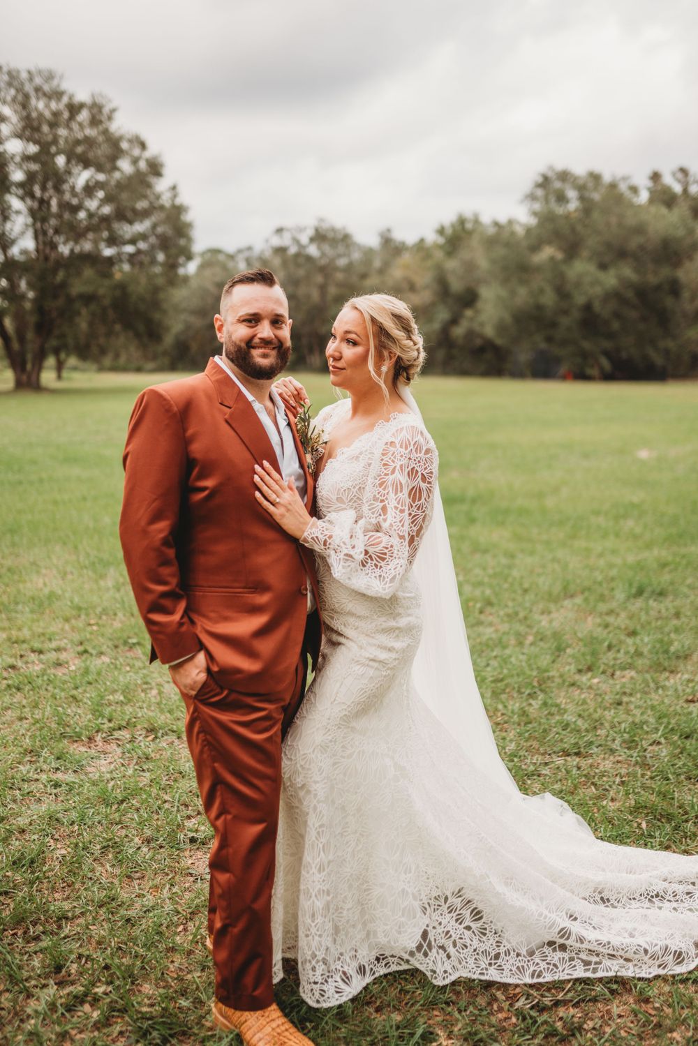 A romantic wedding photo of a couple in a grassy field, groom wearing a rust-colored suit and bride in lace gown.