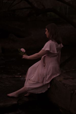 Figure in a pink dress sits on rocks holding a flower in moody lighting.
