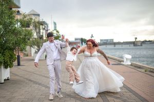 Wedding couple in white attire walking along a waterfront promenade during sunset.