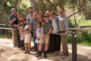 A large multi-generational family group poses together on a wooden railing along a dirt path in a natural outdoor setting.
