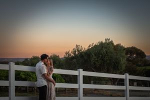 Couple embracing against a white fence at sunset with trees in the background.