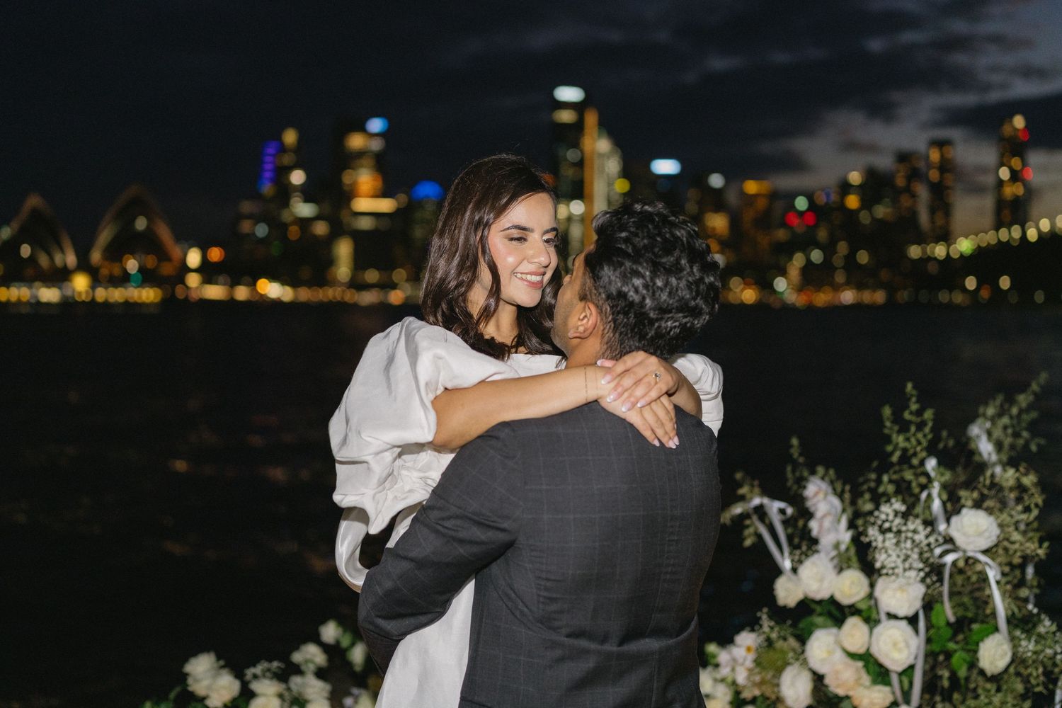 A couple embraces at night with the illuminated Chicago skyline and white floral arrangements visible in the background.