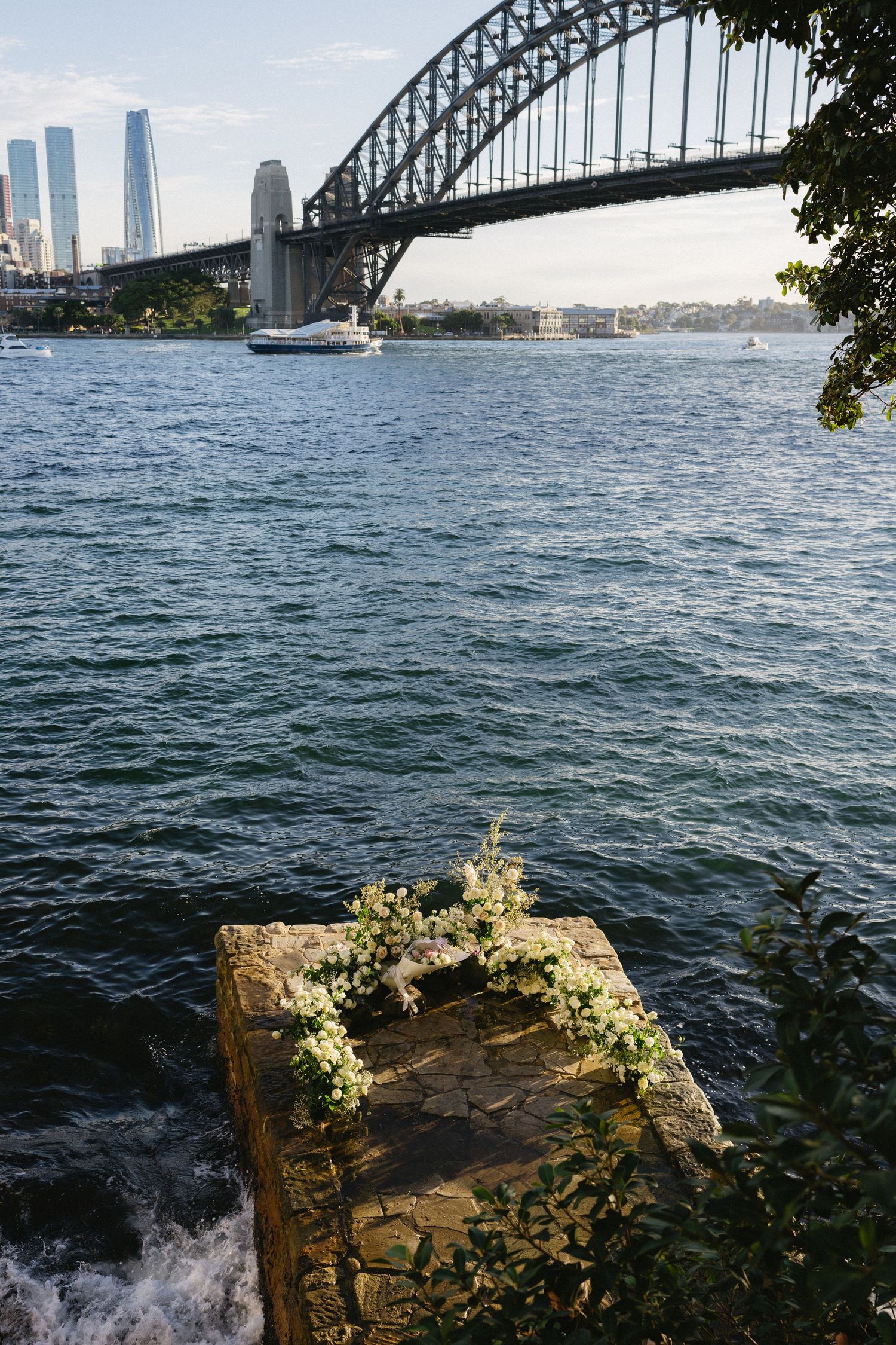 Waves splash against rocky shoreline beneath Sydney Harbour Bridge with city skyline in background.