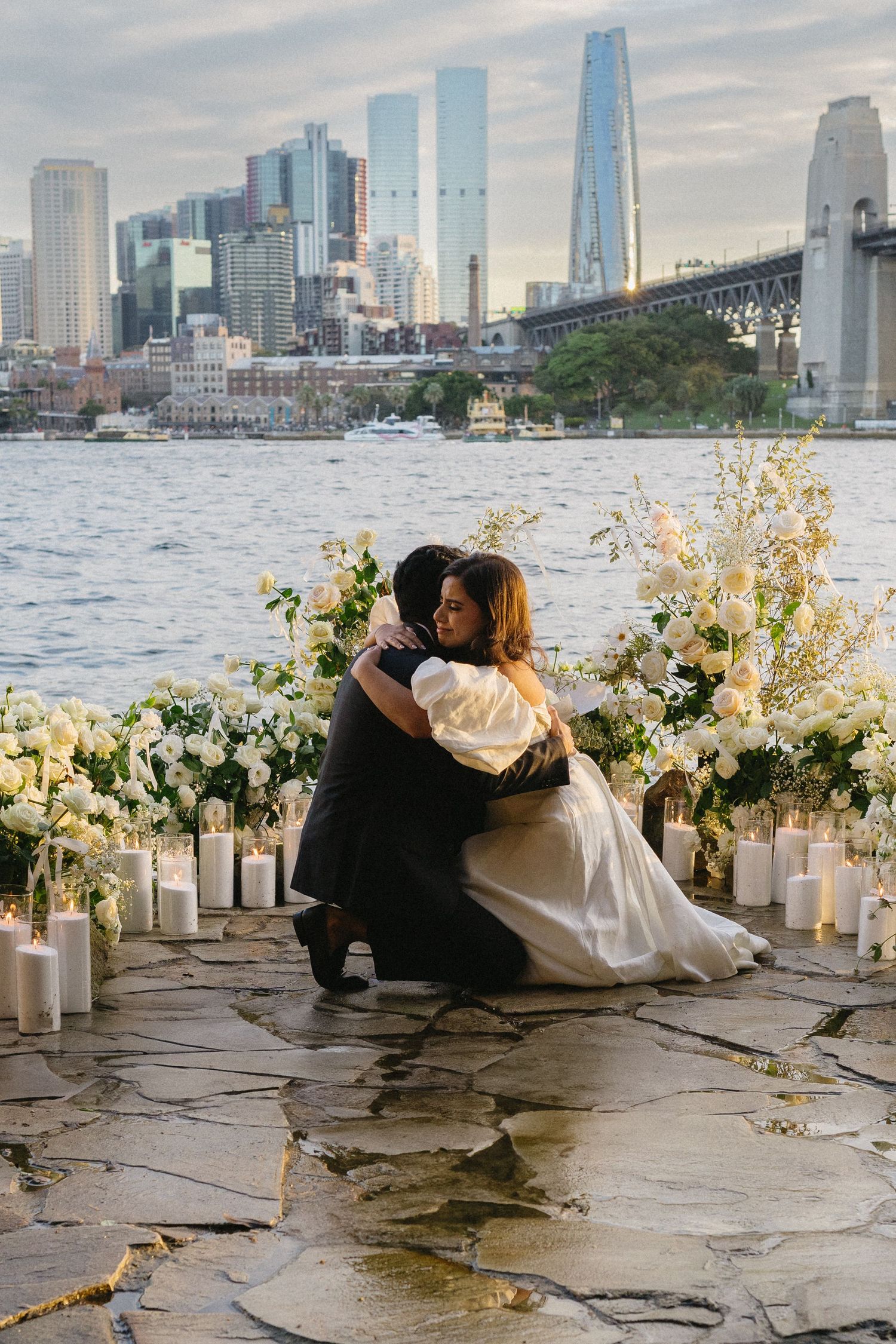 A romantic couple sits by the waterfront with elegant floral arrangements and the New York City skyline in the background.