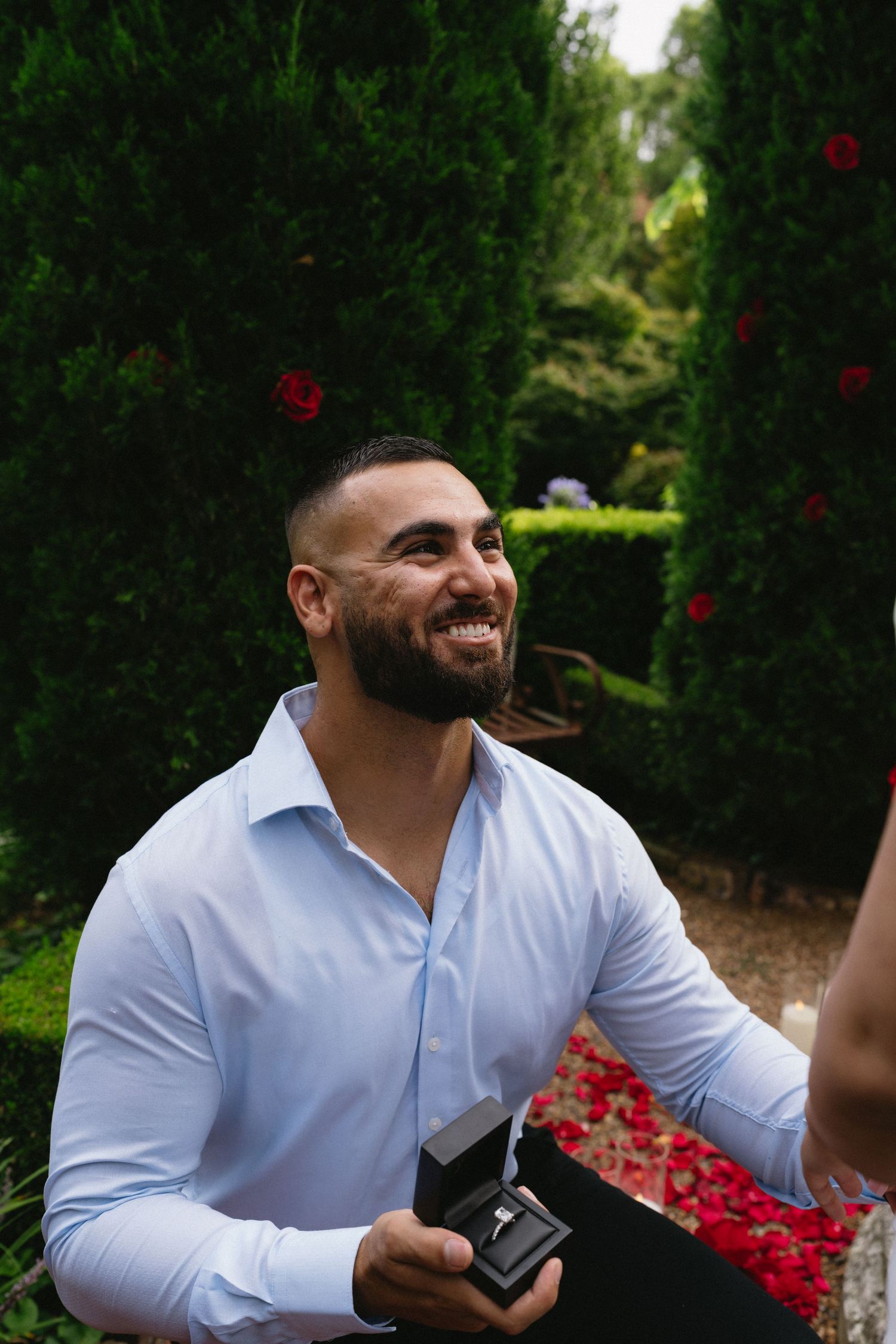 Person in light blue shirt smiling while holding ring box in garden setting.