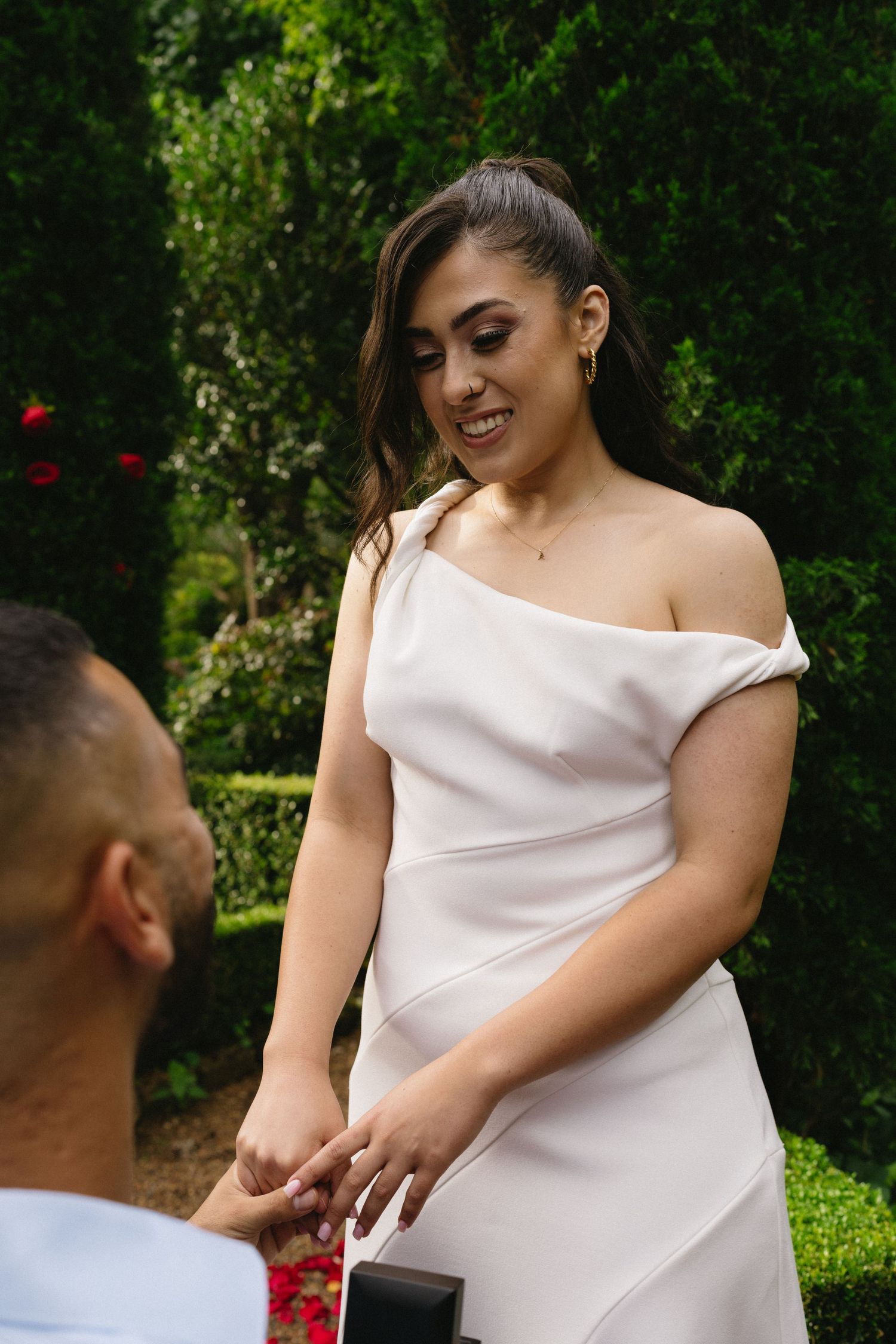 White dress-clad person smiling during romantic garden proposal moment.