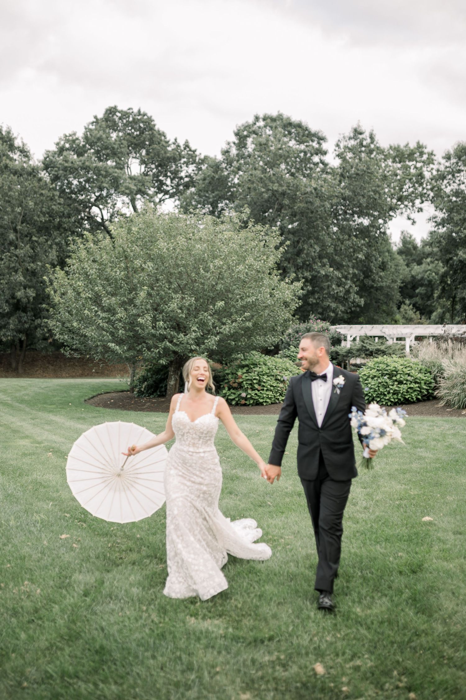 Newlyweds walk hand in hand across lawn with bride holding parasol and bouquet.