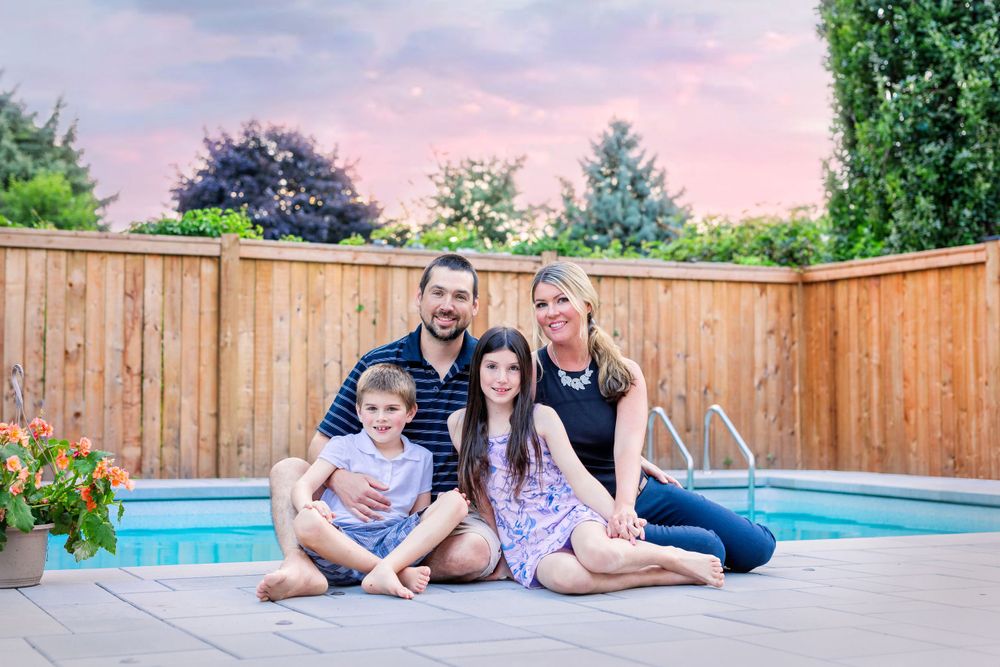 A family sits together on the edge of a backyard pool with a wooden fence during sunset.