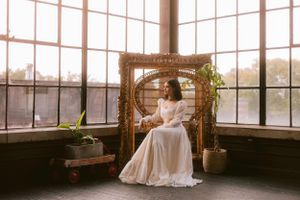A vintage-inspired bridal portrait in a sunlit conservatory with a wicker peacock chair and greenery.