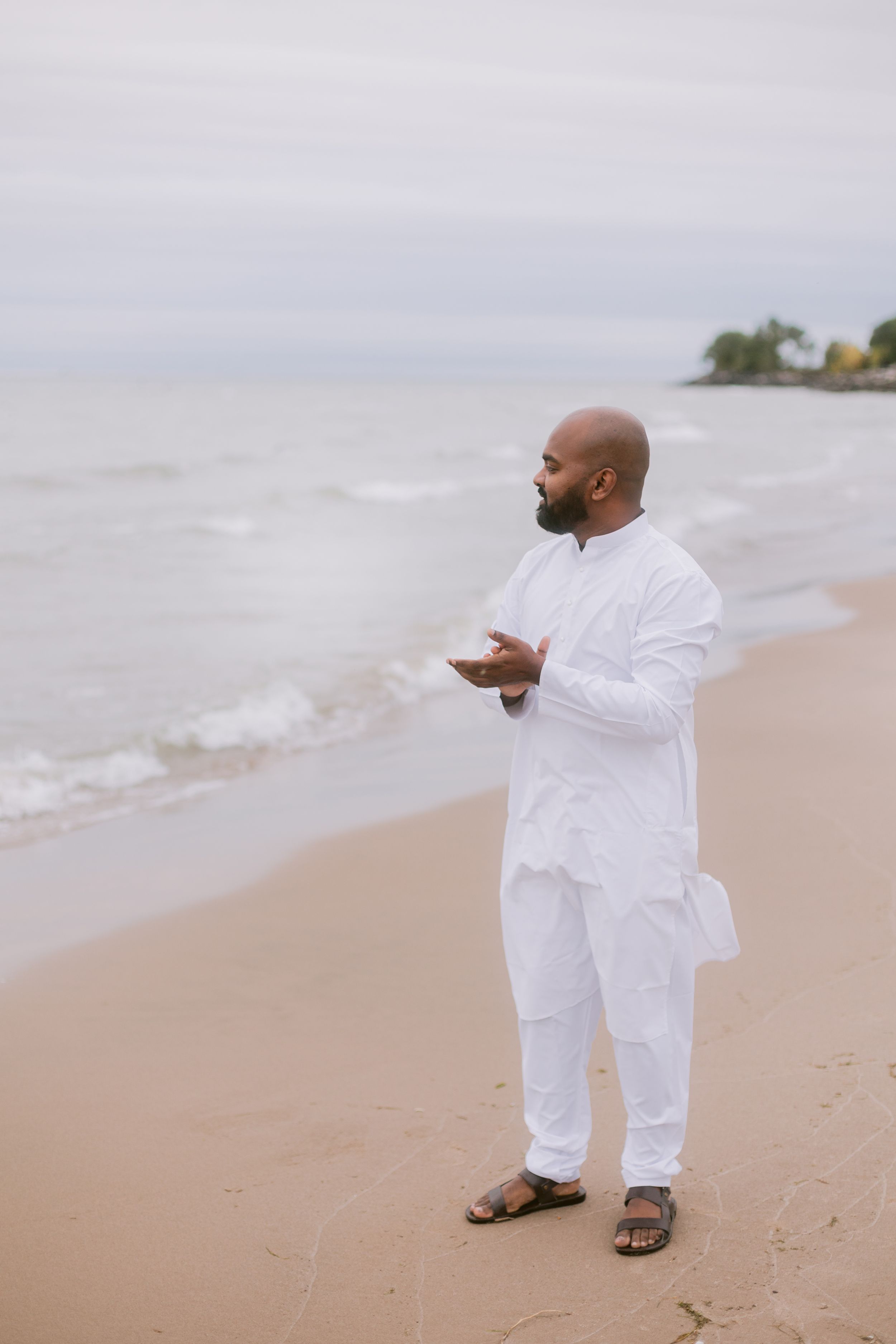A person in all white clothing stands contemplatively on a cloudy beach.