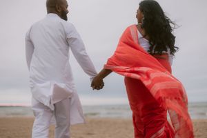 A couple in white and red attire hold hands while walking on a beach at sunset.