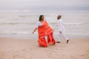 A couple in white and coral outfits dance joyfully on a sandy beach.