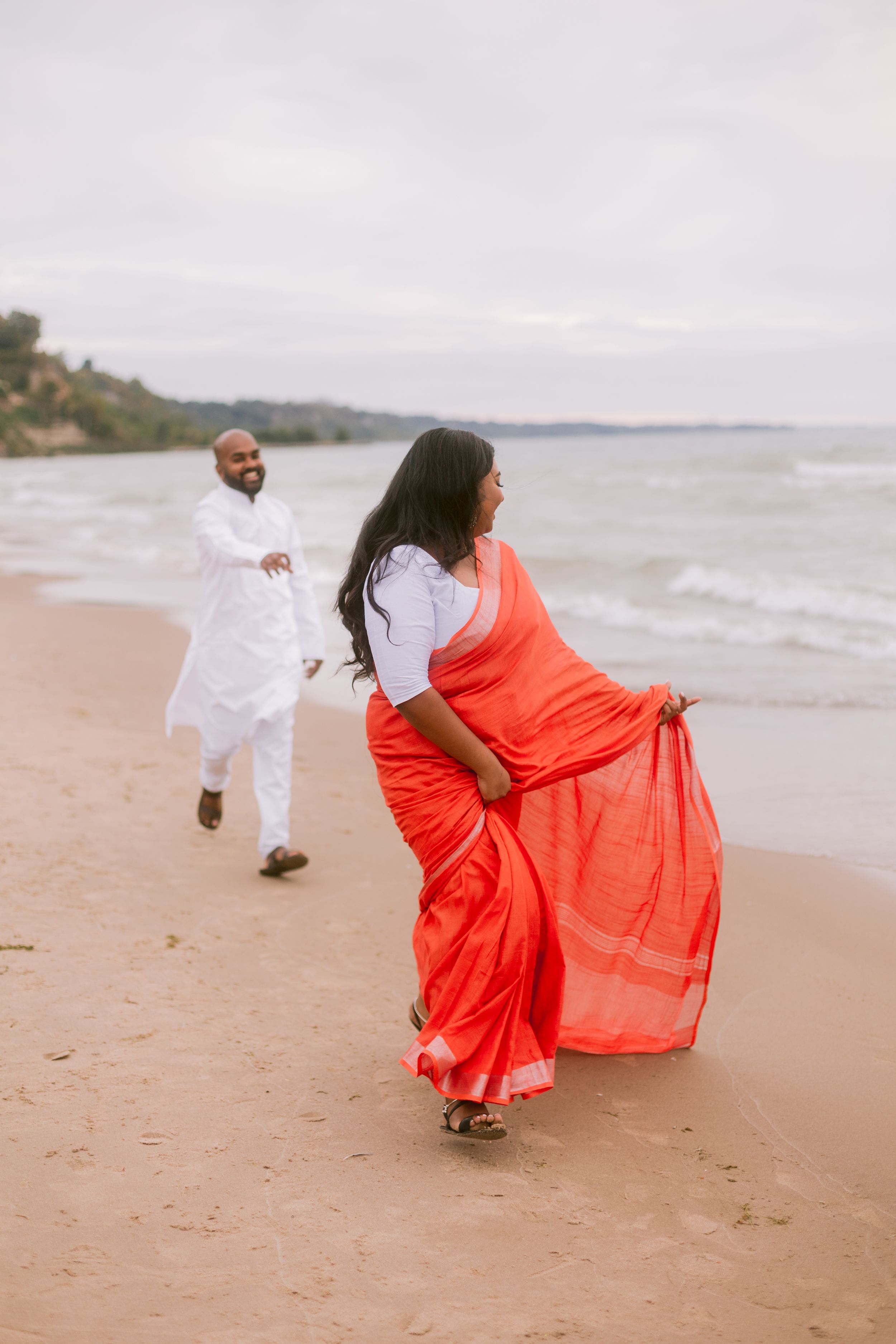 A couple in white and coral attire walk playfully along the shoreline.