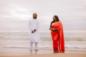 A couple stands on a beach at sunset, with one person wearing white and the other in a vibrant red sari.